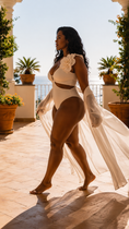 Load image into Gallery viewer, A woman walks barefoot on a sunlit terrace wearing a white one-piece swimsuit with a large floral detail on the chest and a flowing sheer white cover-up draped over her arm.
